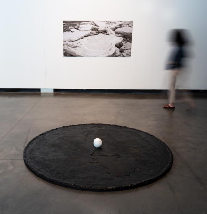 A large photography mounted on the wall. A large circular sculpture made of natural beeswax with a sphere of ice and soda ash melting. A person walks towards the image. 