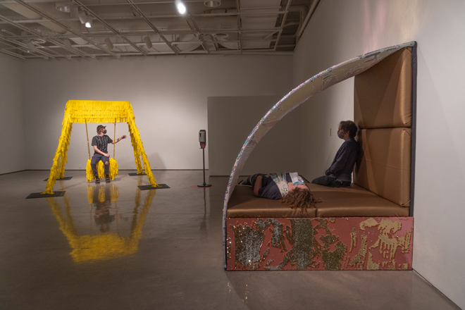 overview of room. Someone interacting with swing set sculpture. Another person inside an enclosed seating