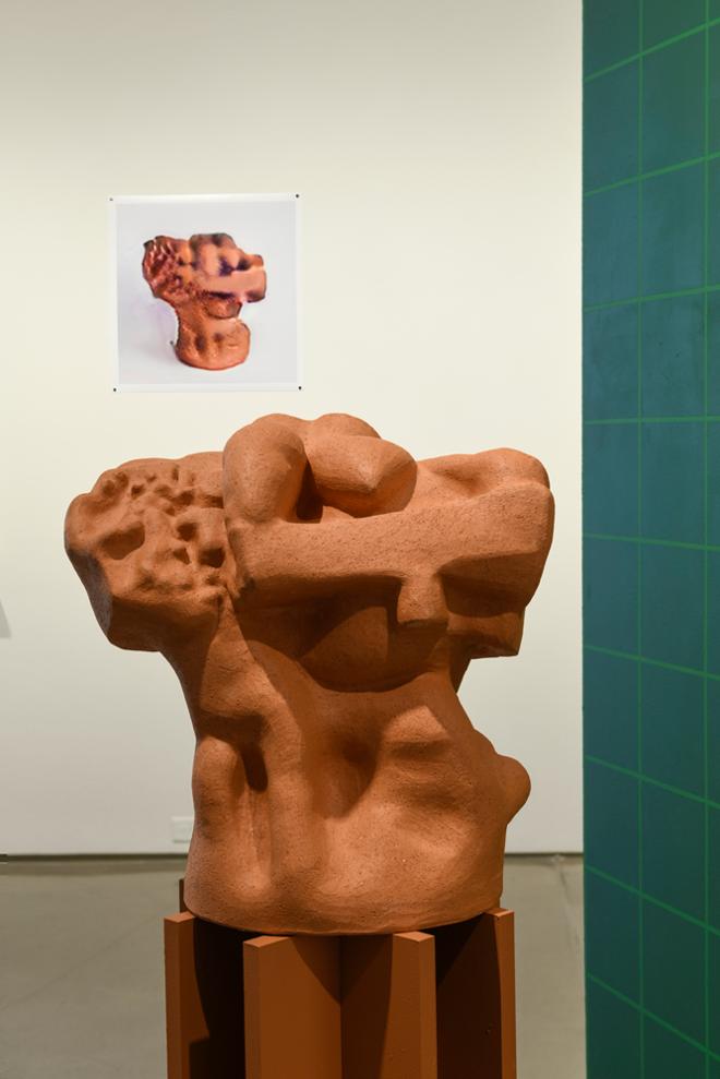A terra cotta sculpture on a color matched pedestal sits in front of a wall mounted image of a computer-generated image of the same 