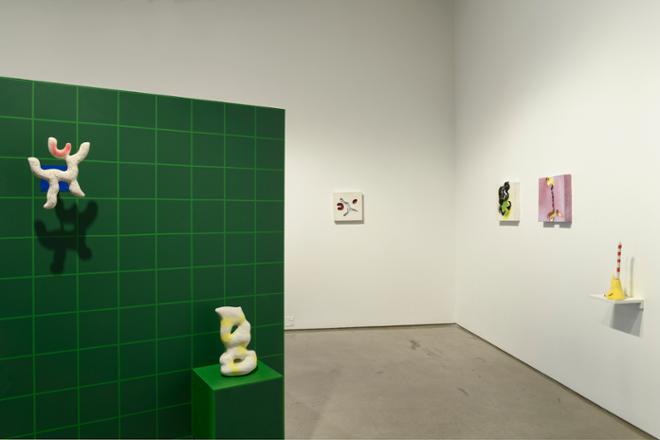 A wide angle shot including a gridded wall with two wall mounted sculptures. Three wall mounted tiles and a small shelf with a sculpture can be seen in the background. 