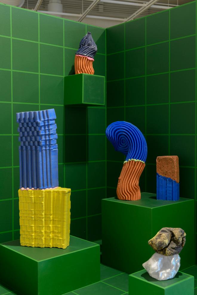 A green, gridded, three-plane display with small cubes seemingly extruding from the surfaces display five colorful sculptures. 