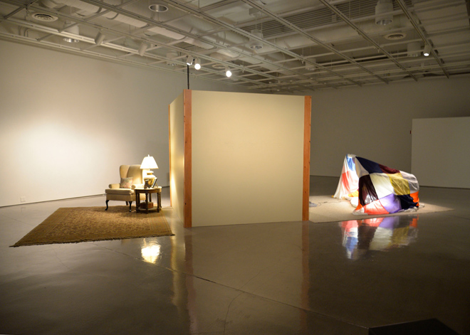 A 8 foot by 8 foot enclosed room with wood trim stands in the askew in center of a gallery. The left wall is the backdrop to a parlor type room with a carpet, armchair and side table. The center wall has three-dimensional text that faintly emerges from the surface. The right wall has a blanket fort containing two neon forms that faces a tube television connected to a video game