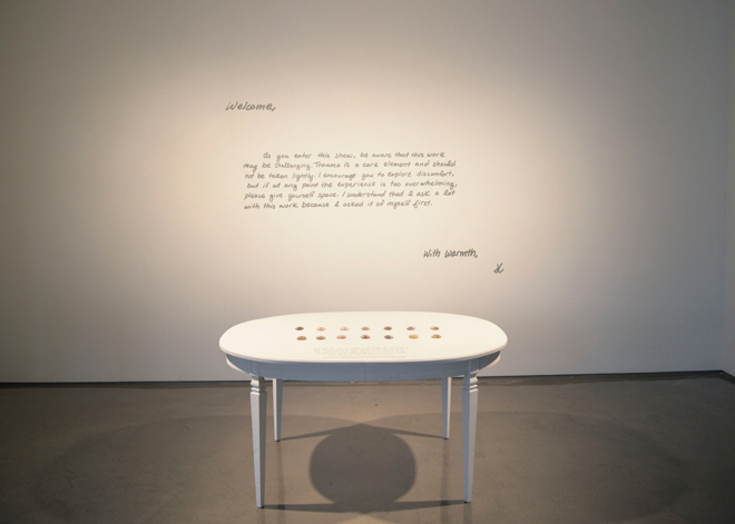 A round, white kitchen table is centered in front of a wall with text on it. The text reads: “Welcome, As you enter this show, be aware that this work may be challenging. Trauma is a core element and should not be taken lightly. I encourage you to explore discomfort, but if at any point the experience is too overwhelming, please give yourself space. I understand that I ask a lot with this work because I asked it of myself first. With Warmth, K” On the kitchen table are fourteen small, colorful glass worry-stones evenly spaced.