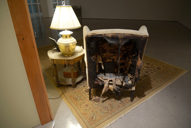 The back of a wing-backed chair that has been burned away. The damage does not extend beyond the back, hiding it when looking at the chair from the back. There are three layers of upholstery can be seen in the damage.