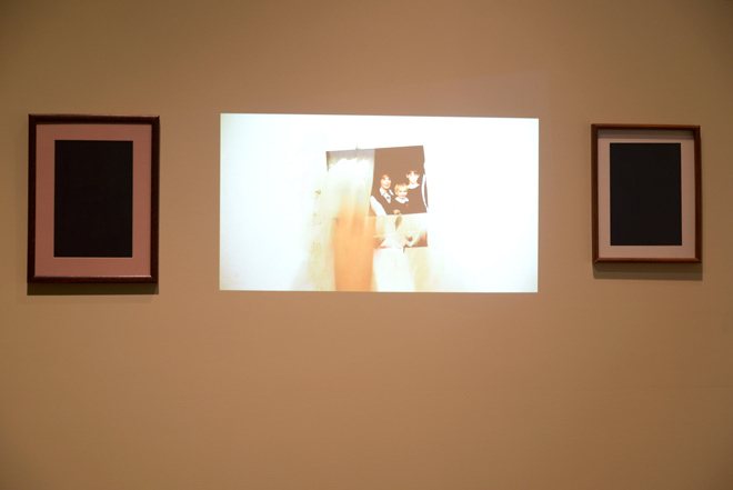Two empty picture frames sit on either side of a life scale projection of a hand patching a hole in drywall. The fist-sized hole is filled in with layers of photographs and spackle.