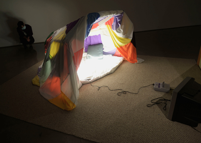A surface of carpet holds a tunnel shaped blanket fort faces a CRT TV connected to a video game system. Inside of the fort, bright light emanates from neon forms. At the back of the fort, a viewer peers through the opening.