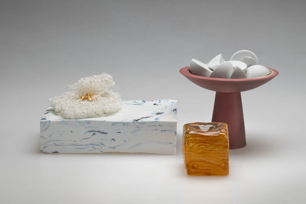 an arrangement of items. To the left features a rectangular object with white and blue marbling. A sponge-like item rests on top. To the right features a plum-colored bowl stand containing white half and quarter circles. A honey-colored, glass square sits in the foreground of the space.  
