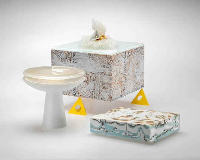  an arrangement of items. In the back of the space sits a white square with wood-texture detailing. A glass slab rests on top of the square, while a sponge-like item sits on top of it. Two small, yellow pyramids sit at the base of the square. A white bowl stand containing a half sphere of glass is to the left of the square. To the right sits a rectangular objects with white, blue, and brown marbling.