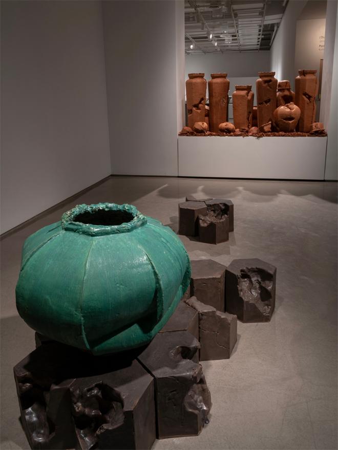 A 2 foot wide bulbous Moon Jar with Green Glaze sits on an assembled honeycomb of hexagonal blocks that look geologically weathered. Behind is a 20 inch tall plinth packed with 38’’ tall urn forms, and an ecosystem of smaller pots, clay chunks, and larger than life seed pods 