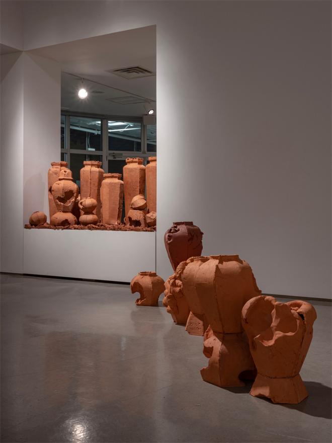 a 20 inch tall plinth packed with 38’’ tall urn forms, and an ecosystem of smaller pots, clay chunks, and larger than life seed pods, in the foreground, ginger jar forms are emerging from the gallery floor, having been bifurcated horizontally