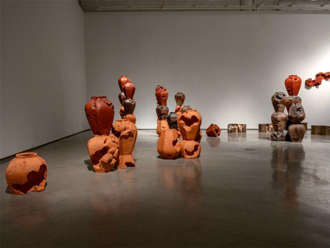 A sprawling arrangement of ginger jars in terra cotta red, multi-shade brown, and bright bisque orange rise into the distance.  They are stacked into columns, and vary in height.  Their placement follows a meandering line, like the ridge of a mountain range or the path of a creek