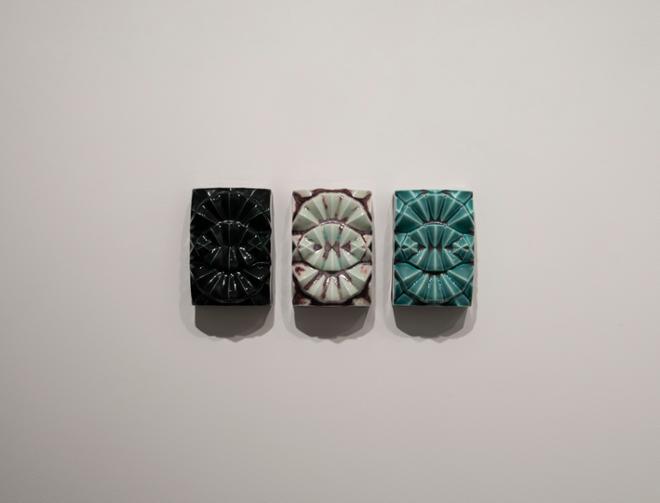 three small, rectangular tile pieces arranged next to each other on the wall. Features a shell-like design, the one of the left black, the one in the middle light blue with plum detailing, and the one on the right teal