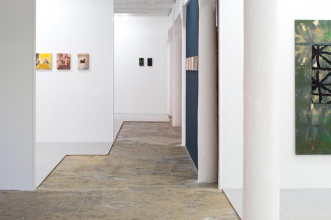 An installation shot looking through the walkway. To the right is a part of the painting Untitled (Green and Orange). Looking further is the installation room of Isabel Monti with multiple miniature paintings on white wall (to the left) and dark blue wall (in the middle). At the end of the walkway is Untitled #8 and #11.  