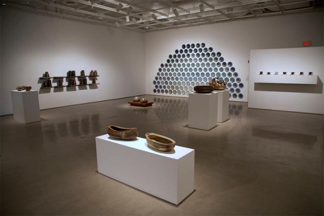 Wide-angle view of Jake’s show in Fosdick-Nelson Gallery, Time is a Flat Circle. In the foreground, there are several wood fired vessels on pedestals. On the left side of the gallery there is a grid of shelves on the wall, five across and two deep, with jars sitting on them, called Markers of a Place and Time. On the back wall is an installation of 158 plates, called Big Sky, which are glazed in a gradient of blue. On the right side of the gallery is a display of cups on a shelf. 