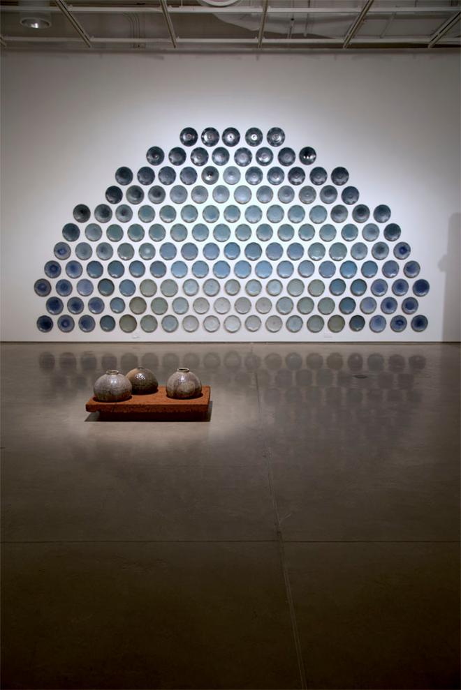 Installation of 158 plates hanging on the wall which are glazed in a gradient of blue from light in the bottom-center to dark on top. Each plate is roughly 10” wide. In front of the wall is a clay plinth on the floor of the gallery that is approximately 3.5’ wide, with three low, bulbous vessels on top of it. 