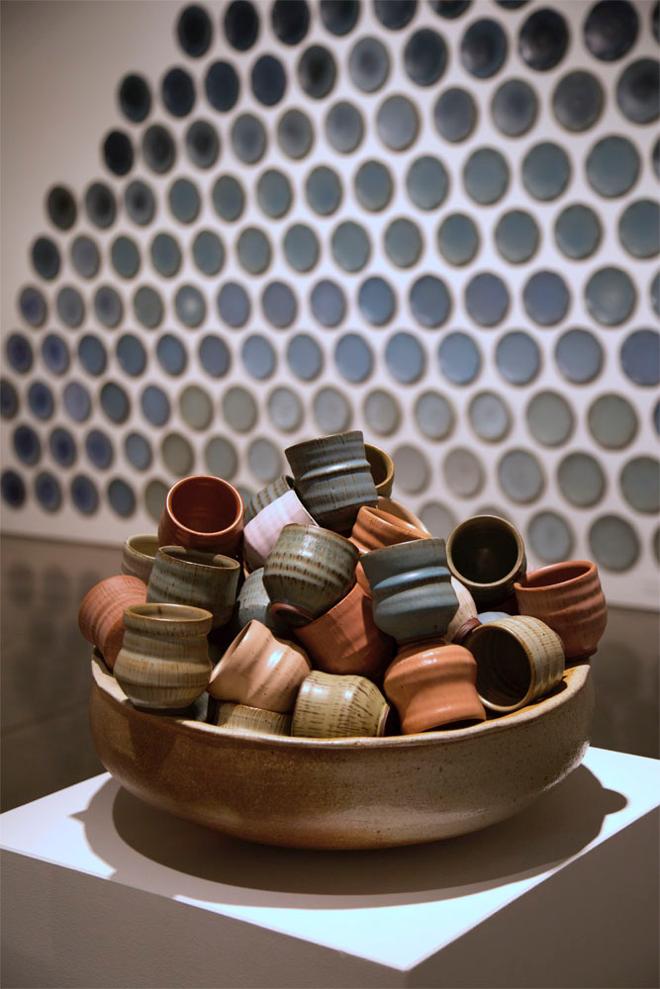A large bowl piled full of cups sits on a pedestal, with a wall of plates behind it. The cups are a range of shapes and are glazed in a variety of colors with satiny glazes. It feels like a cornucopia of pots that you want to explore and pick through, but they are piled rather precariously in a way that makes you nervous to touch them. 