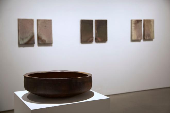 A large, dark basin sits on a pedestal in front of a wall of six large ceramic tiles. You can’t see much of the interior of the basin, which is in shadow, but can see the profile and the shadow cast underneath it. The tiles, which are out of focus in the background, are each 20” tall and are hanging on the wall in three mirrored pairs. The outer pairs are lighter colored and the middle one is darker.  