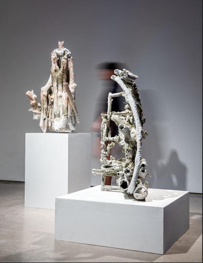 Installation image of two sculptures on pedestals. A blurry human visitor is seen in the background. One of the sculptures consists of vertically arranged hollow branches, glazed in white and pink tones. The other sculpture is a algae -green grid. Fabrics and hollow branches are interlaced with the grid. 