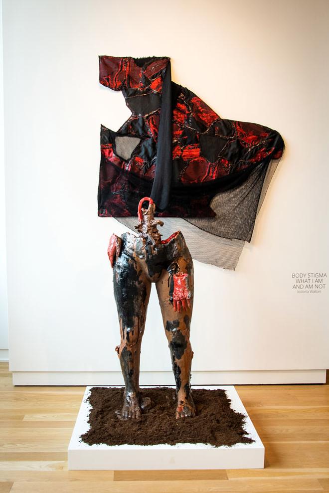 The figure is partial from the feet to mid-torso. Two red hands are attached around the waist, displaying different qualities of pain, one through a bubbly surface and the other through engaging with the wound on the side of its body. Various glazes and spray paint are splashed on the brown skin, like tar. Soil fills the vessel of the body and surrounds the piece.  