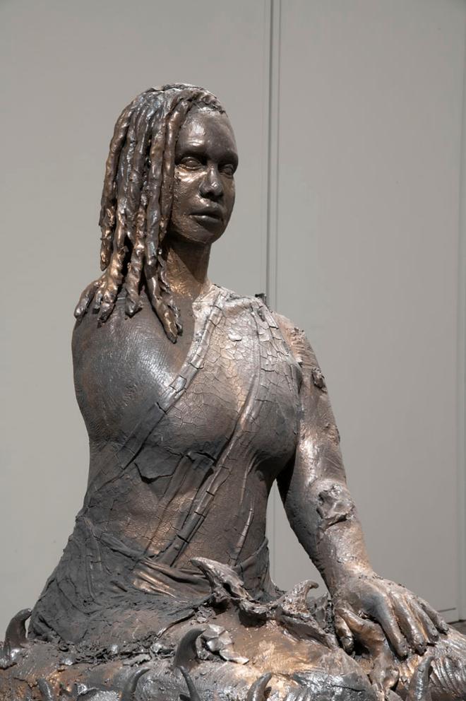 Like A Thorn is a ceramic figure of a Black woman who is an amputee, seated with their legs and the surrounding base as a bed of thorns. Her pose is relaxed and her gaze is fixed ahead. She is wearing a flowing dress, caked and cracking like dried earth. Her skin and surroundings fluctuate between golden bronze, dark brown, and black.  