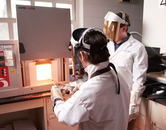 Students pulling molten glass from a furnace wearing gloves and other protective gear