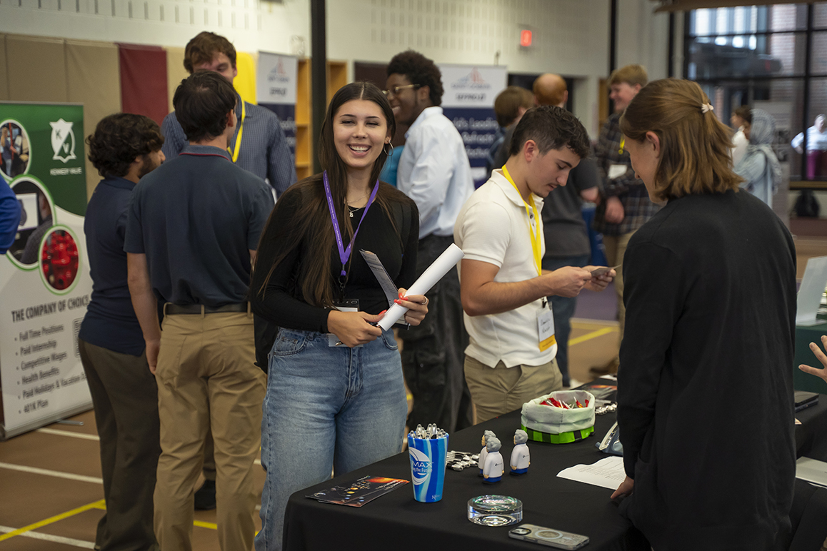 An Alfred University student smiles while holding papers and standing at an employer table during the campus Career Fair.