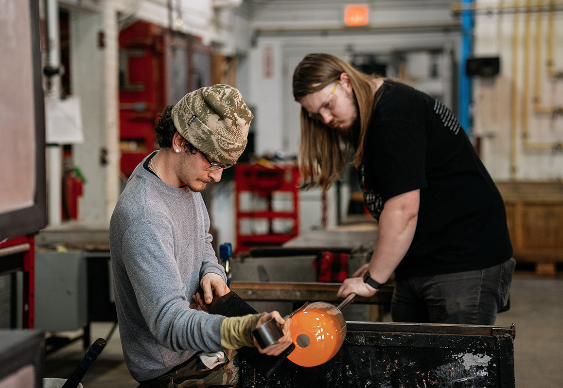 A photo of two Alfred University students shape hot glass