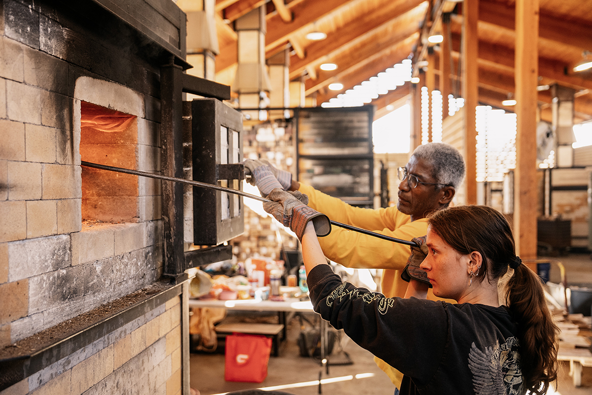 A photo of an Alfred University student and faculty member reaching into a kiln