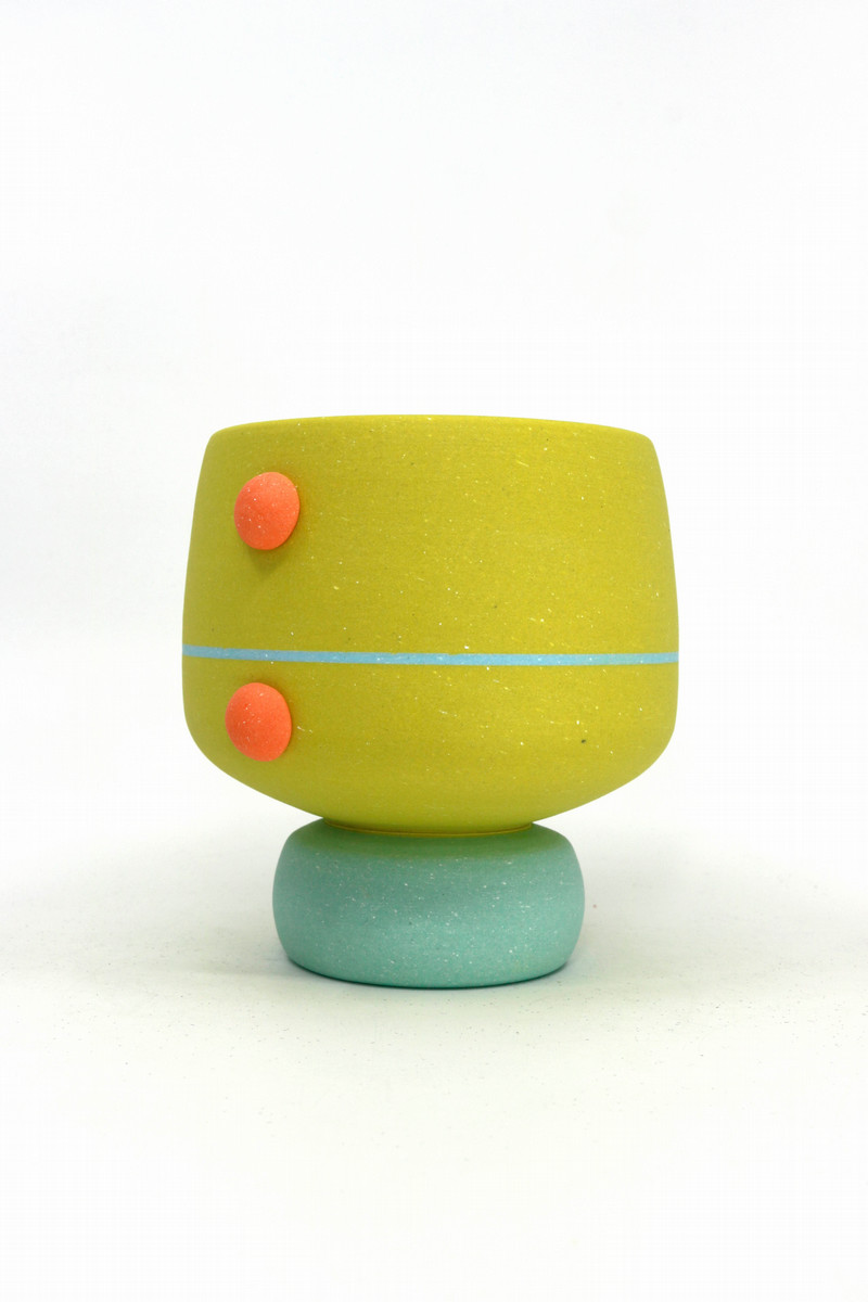 Modern ceramic cup with a yellow body, accented by two small orange buttons on the side, a thin blue horizontal stripe around the middle, and a round light blue base. The design is simple and playful, emphasizing contemporary style.