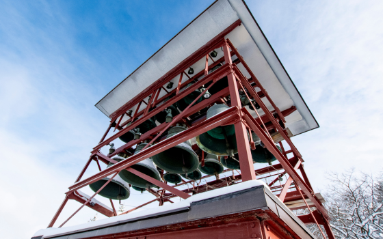 Red metal bell tower with green bells, viewed from below against a bright blue sky. Snow rests on the structure, conveying a crisp winter atmosphere.