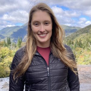 A woman with long brown hair stands outdoors, smiling. She wears a black jacket. Behind her are lush green mountains and a partly cloudy sky.