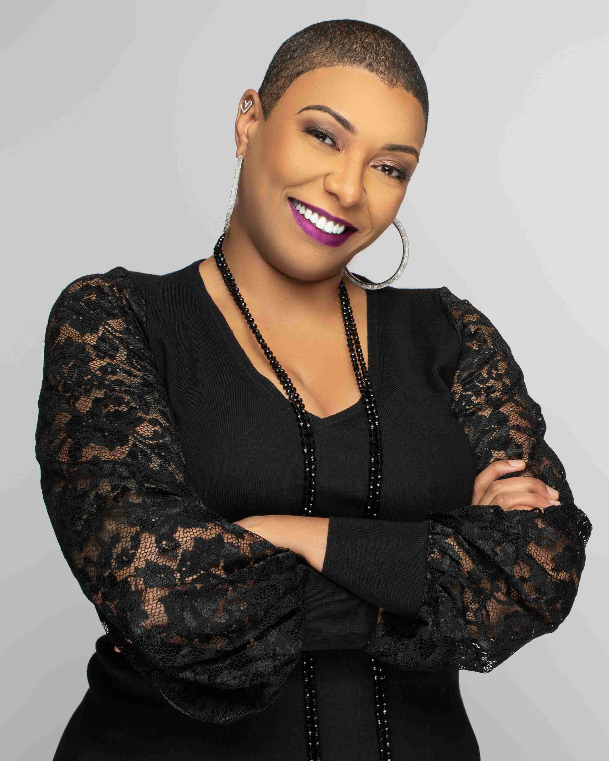 Smiling woman with short hair in a black dress with lace sleeves, wearing hoop earrings and a necklace, arms crossed, exuding confidence.