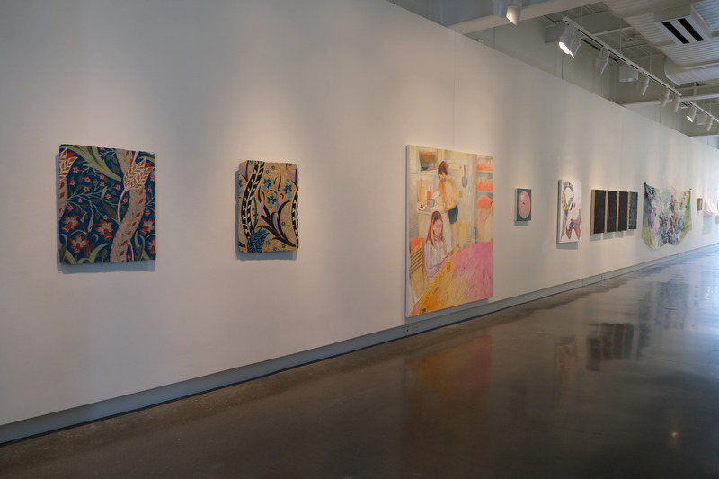 Gallery wall featuring a range of contemporary artworks, including botanical illustrations, figurative scenes, abstract compositions, and a grid of monochrome canvases. The space is illuminated with overhead lights, highlighting the colorful and textured pieces along a white-painted wall with a polished concrete floor.