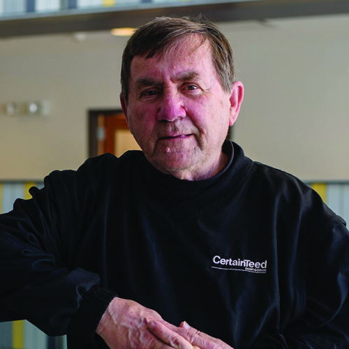 A man in a black CertainTeed sweatshirt stands indoors, leaning casually on a surface. The setting is softly lit, conveying a warm tone.