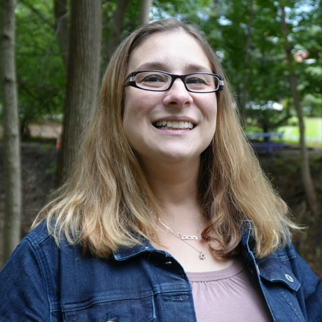 A smiling woman with long light-brown hair and glasses stands outdoors in a wooded area. She is wearing a denim jacket over a light pink top. Trees and greenery fill the background.