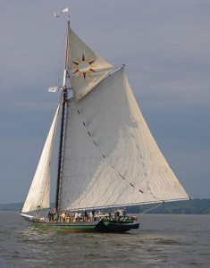 A large sailboat with a tall mast and white sails glides on open water, under a cloudy sky. A sunburst pattern decorates the sail. People are on deck.