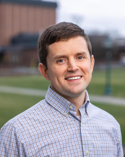 A smiling man in a plaid shirt stands outdoors. The background shows a blurred campus with green lawns and a brick building, conveying a cheerful, academic setting.