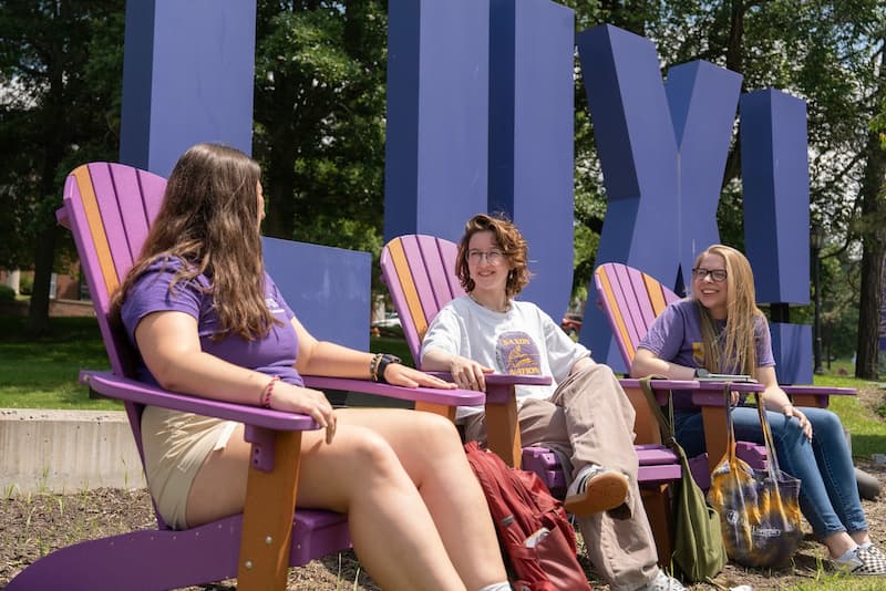 Three Alfred University students chat and relax in purple Adirondack chairs in front of the large AU letters on campus.