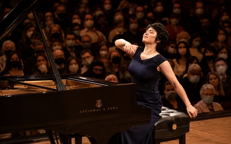 A young woman in a navy blue dress plays a Steinway and Sons grand piano with her eyes closed and arms extended, performing passionately before a live audience.