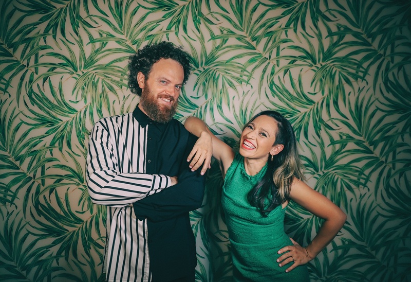A man with curly hair and a beard wearing a black and white striped shirt and a woman with dark hair wearing a green dress smile and laugh together in front of a tropical leaf wallpaper background.