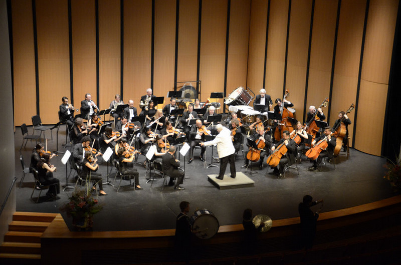 A full orchestra dressed in formal black attire performs on stage in a concert hall, led by a conductor in a white jacket standing at the center podium.