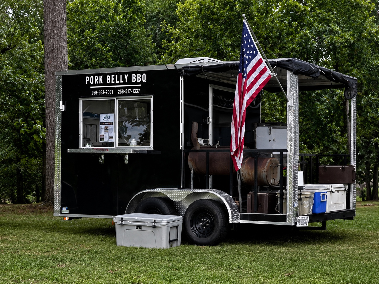 The Pork Belly BBQ food truck trailer set up outdoors with a smoker and an American flag displayed.