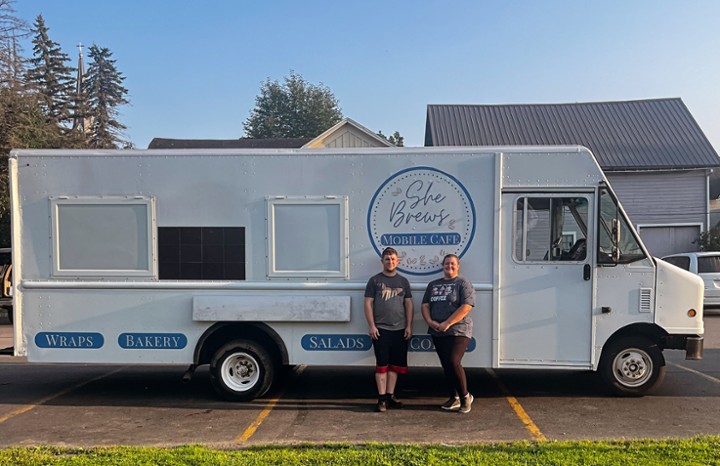 Two staff members stand in front of the She Brews Mobile Café truck, which displays wraps, bakery, and salads on the side.