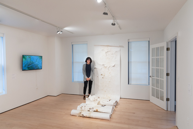maria posing next to her artwork of a long scrolling material hung on the wall and draping along the floor