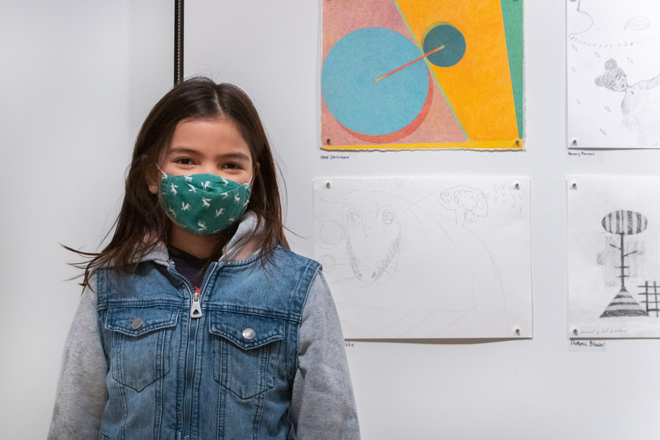 little girl in front of artwork wearing a green face mask smiling