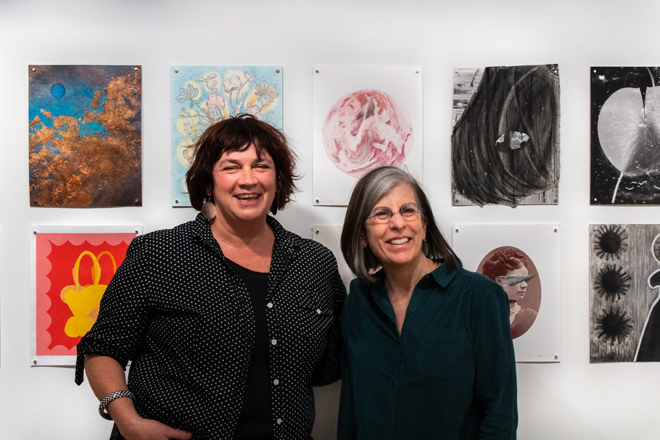 Coral and Sharon posing in front of drawings on display and smiling