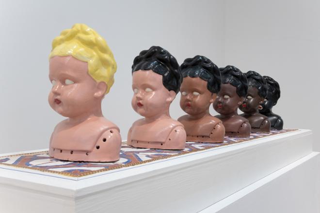 series of doll like sculpture heads in various hair and skin tones in order from lighter features to darker features