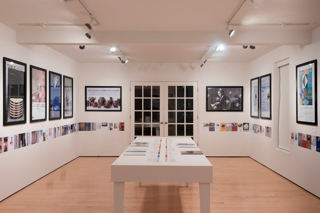 Wide-angle view of the Cohen Gallery during the Poster Archives 2001–2024 exhibit. The room features framed posters along all three walls and a white display table in the center with open catalog pages and colored timelines. The lighting highlights the clean, minimalist design of the space.