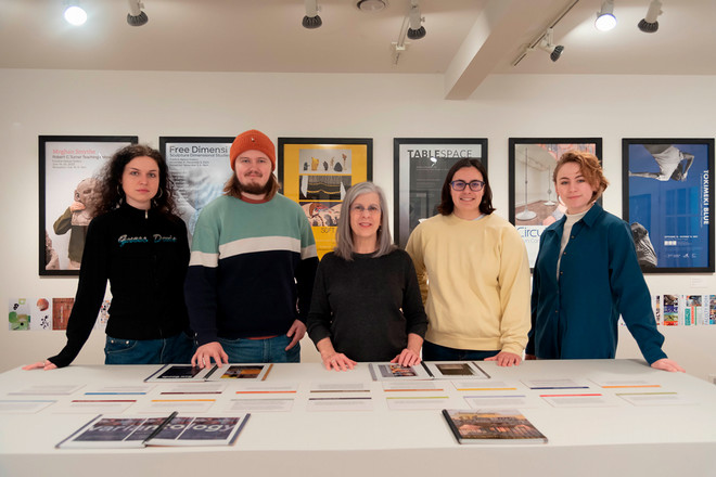 A group portrait of four gallery interns standing alongside Gallery Director Sharon McConnell at the center of the Cohen Gallery. They stand behind a table displaying the exhibition catalogue and printed materials, with framed posters from the Poster Archives exhibit on the wall behind them.