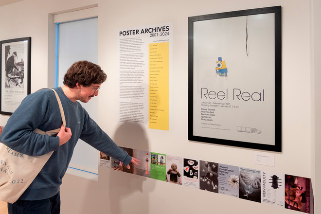 A gallery visitor wearing a blue sweatshirt and tote bag points at a row of small posters beneath a framed exhibition print titled “Reel Real.” Next to it is a large text panel titled “Poster Archives 2001–2024,” detailing the names and history of the internship and exhibit contributors.